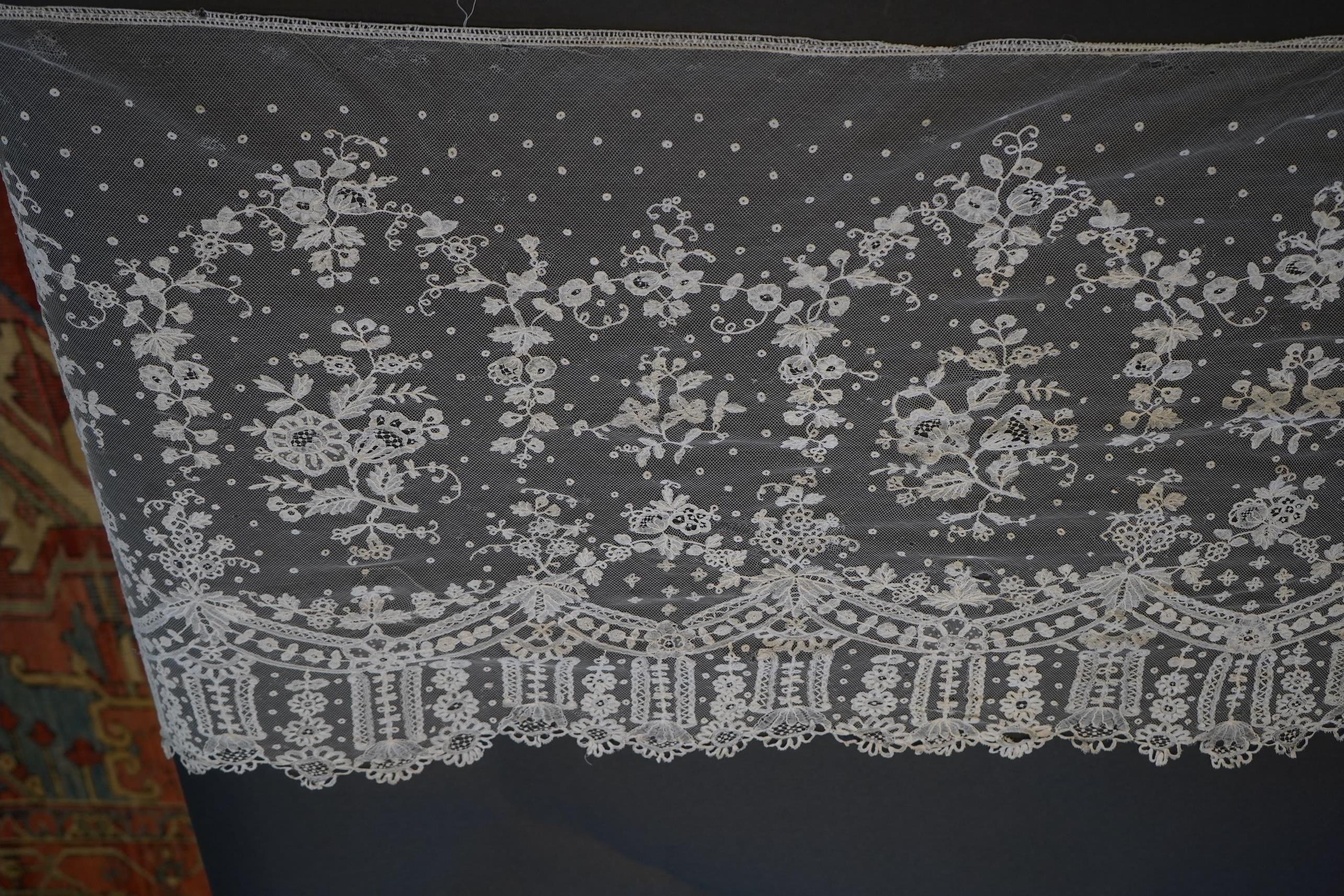 A deep elaborately designed flounce of mid 19th century Brussels bobbin appliquéd lace on net, with finished lace ends, worked with an intricate floral border below large floral cartouches framed in similar motifs and al